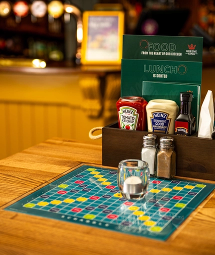 A close up view of menus, cutlery, condiments, and a tea light sitting on a wooden restaurant table at the Malvern Tavern. A Scrabble board is inlaid into the table.