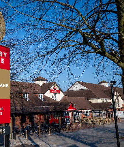 A close up view of the exterior sign of The Weathervane and the trees surrounding the building, with the pub visible in the background.