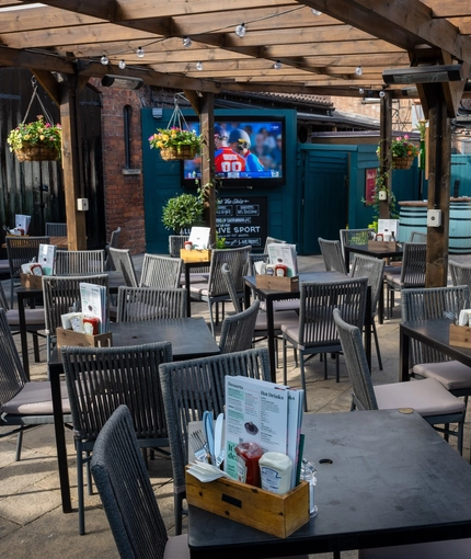The paved, exterior beer garden seating area at The Ship; with a large wooden pergola sheltering the tables and chairs, flower baskets and string lights hanging from the dark wooden beams, and a TV on the wall.