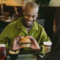 Person seated at a pub table holding half of a vegetable burger, with the other half and a portion of chips on the plate, alongside a glass of red Old Mout cider and a pint of Peroni.
