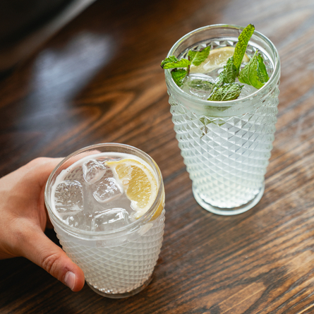 Two cocktails served in glasses which sit on a wooden table. Both cocktails are served with ice and slices of lemon, one is also garnished with mint, and a hand is holding the other cocktail.