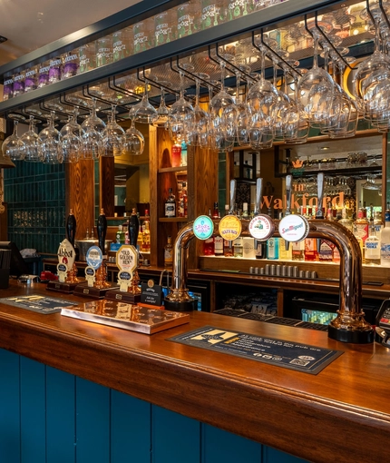 A close up view of the bar inside The Walkford in Walkford, with glasses hanging in racks above the counter, and bottles of alcoholic spirits on wooden shelves behind the bar.