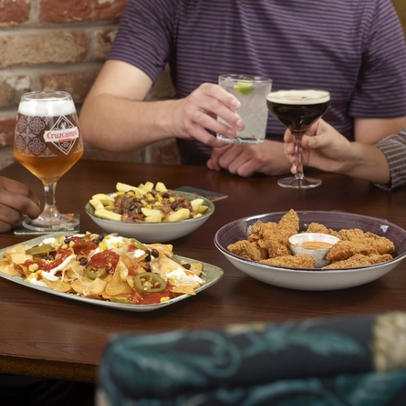 A table with sharing plates of food, including nachos, loaded fries, and breaded bites with dipping sauce, while several people seated around the table hold various drinks such as beer and cocktails.