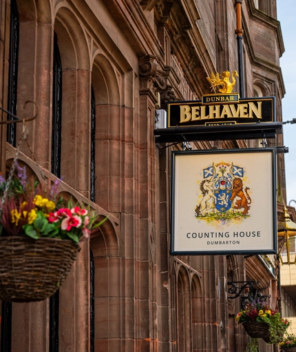 A close up view of the pub sign on the exterior wall of the Counting House. The sign features an illustration of a coat of arms held by a unicorn and a lion, above the words "Counting House, Dumbarton", on a white background.