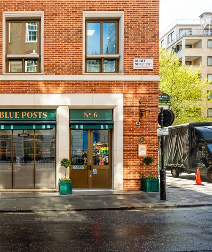 The exterior facade and signage of the Blue Posts in St James's, with small trees in green planter boxes on each side of the double doors.
