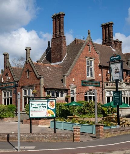 The exterior facade, signage, and beer garden seating area of the Golden Hind in Cambridge, with wooden picnic tables, shade umbrellas, and string lights.
