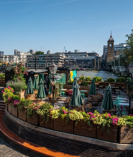 The exterior beer garden seating area at The Anchor Bankside in Southwark; with shade umbrellas on some of the tables; string lights hanging overhead; a view of the River Thames; and steps leading up to the seating area which is surrounded by planter boxes filled with flowers. 