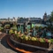 The exterior beer garden seating area at The Anchor Bankside in Southwark; with shade umbrellas on some of the tables; string lights hanging overhead; a view of the River Thames; and steps leading up to the seating area which is surrounded by planter boxes filled with flowers. 