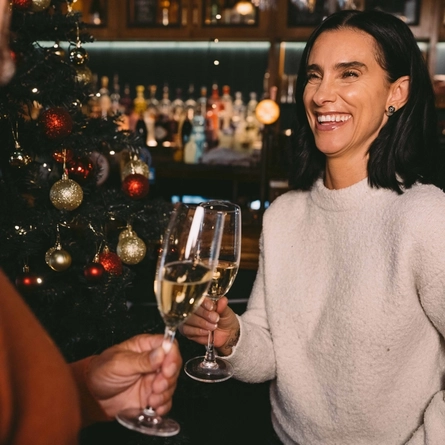 Two people holding glasses of sparkling wine, standing in a bar next to a Christmas tree decorated in red and gold.