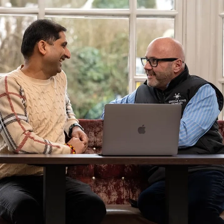 Two Men sat on a sofa facing each other and smiling, there is a laptop on the table in front of them. 