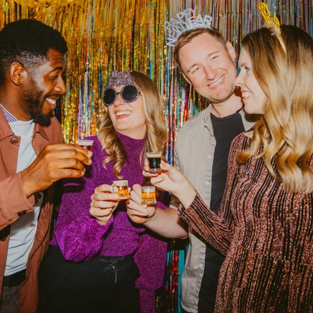 Four adults celebrate New Year’s Eve in front of a shimmering gold and silver tinsel backdrop, holding miniature beers and layered shot glasses. Several people wear novelty “Happy New Year” headbands and oversized party glasses, while glittering outfits and sparkling decorations create a lively festive atmosphere.