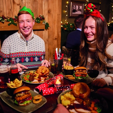 A group of people are gathered around a table enjoying a festive meal. They are wearing paper crowns, and the table is set with plates of food. The background is decorated with Christmas decorations.