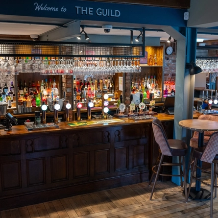 The wood panelled bar inside The Guild, with a table and chairs next to the counter and glasses hanging in racks above the bar.