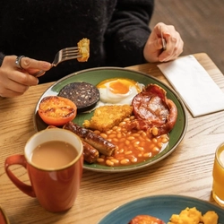 Two plates of Full English Breakfast featuring hash browns, tomatoes, eggs, sausages, bacon, black pudding, and baked beans. The plates sit on a wooden table along with napkins, a cup of tea, and a glass of juice, and a person at the table is holding a piece of hash brown on their fork.