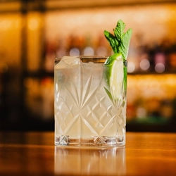 A close up view of a Sparkling Coconut Margarita served in a carved tumbler glass with ice, a sprig of mint, and a wedge of lime. The glass sits on a wooden table in a bar.