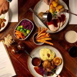 An overhead shot of a range of plated Christmas mains on a festive decorated wooden table, with hands of a person seated around the table holding a knife and fork and another pouring gravy from a mini pan.