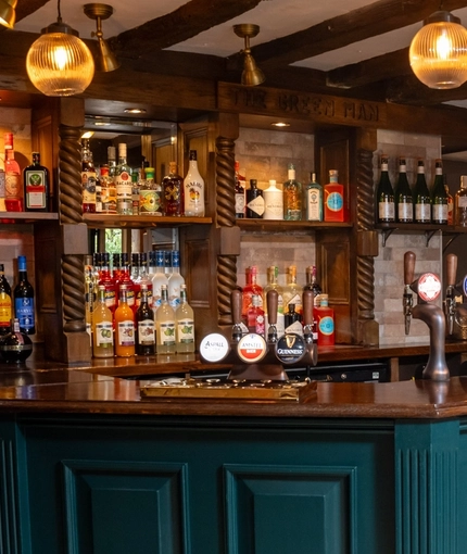 The dark green, wood panelled bar at The Green Man in Great Offley, with stone flooring, wooden beams on the ceiling, and bottles of wine and alcoholic spirits on wooden shelves behind the counter.