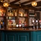 The dark green, wood panelled bar at The Green Man in Great Offley, with stone flooring, wooden beams on the ceiling, and bottles of wine and alcoholic spirits on wooden shelves behind the counter.