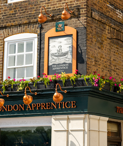 A close up view of the exterior facade and signage of The London Apprentice, with planter boxes filled with flowers above the golden yellow lettering spelling "The London Apprentice" on two sides of the pub.