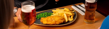 A hand squeezing a lemon wedge over a plated portion of fish and chips, at an indoor table.