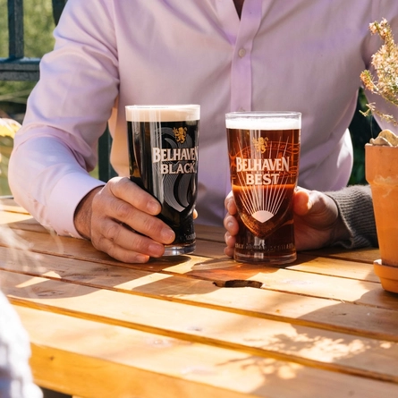 A close up view of two hands holding glasses of Belhaven Black and Belhaven Best, at a wooden table in a sunny beer garden.