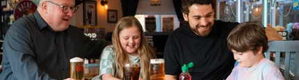 Two adults and two children seated at a wooden table in a pub, looking at colourful activity sheets with crayons. Drinks on the table include two pints of beer, a glass of cola, and a small fruit drink