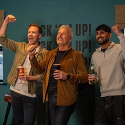 Three people standing in the gaming area of a restaurant, all of them raising a hand and cheering. One of them is holding a glass of beer, one is holding a glass of Guinness, and the other is holding a bottle of Peroni.