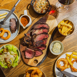 An advertising image showing a boarded Ribeye Sharer dish including onions rings, chips, salad and sauce sat on a table within the interior restaurant seating area at The Four Oaks.