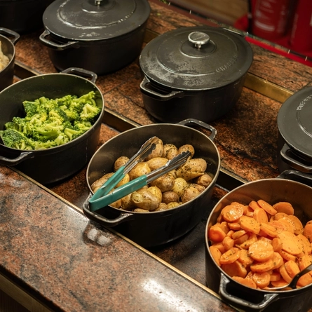Cauliflower cheese, broccoli, new potatoes, and carrots, each served in a large roasting dish which sits on a carvery counter.