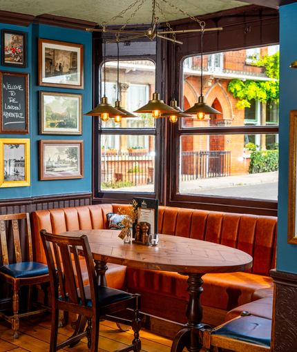 An interior restaurant seating area at The London Apprentice, with a U-shaped booth seat beside the window, framed artwork on the walls, wooden flooring, and a water station.