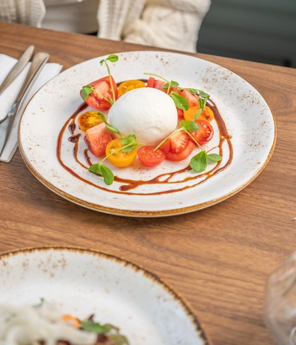A close up view of a Burrata served on a plate with sliced red and orange tomatoes, and drizzled with sauce. The plate sits on a wooden table along with cutlery, a serviette, a glass of white wine, and another plate of food; and a person sitting at the table is picking up the glass of wine.