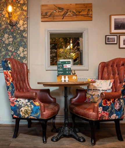 A close up view of one of the small wooden restaurant tables inside The Royal Oak, with two wingback chairs, and framed artwork on the wall.
