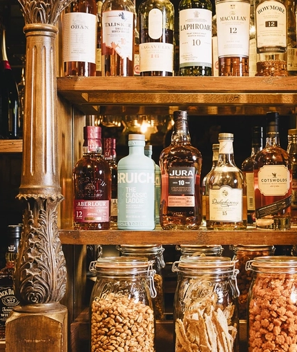 A close up view of bottles of wine, whiskey, and other alcoholic spirits; glass jars filled with nuts and other bar snacks; and glasses, sitting on wooden shelves at the Masons Arms.