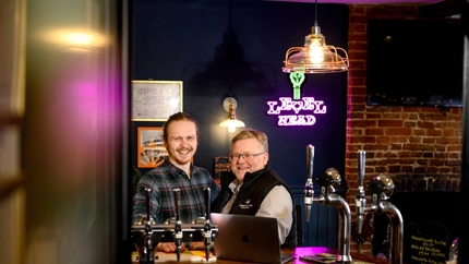 Two Men stood at a bar smiling with a laptop in front of them