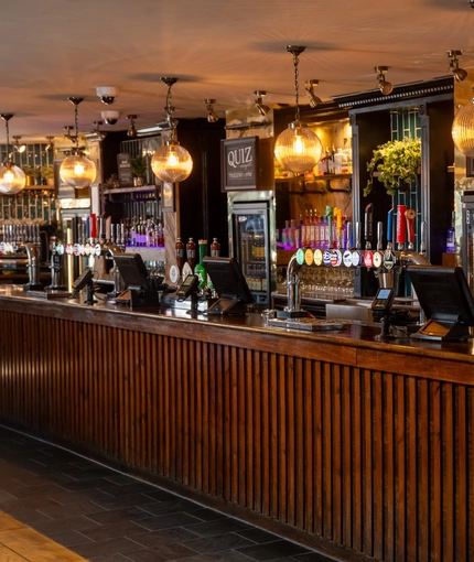 A close up view of the wood panelled bar at the Rudds Arms in Marton-In-Cleveland; with spherical lights hanging above the counter, and bottles of alcoholic spirits on illuminated wooden shelves behind the bar.
