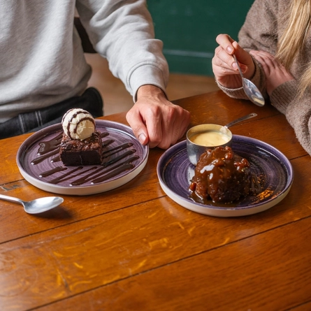 A plate of Sticky Toffee Pudding with a pot of custard, and a plate of Brownie topped with a scoop of ice cream and drizzled with sauce, sitting on a wooden restaurant table. Two people are sitting at the table, one holding the plate of Brownie, and the other holding a spoon.