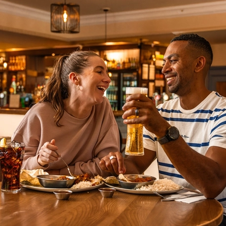 Two people seated at a wooden table in a pub, enjoying curry, rice and naan bread. One person holds a pint of lager while a glass of cola with ice and lemon sits nearby. Warm lighting, exposed brickwork and a stocked bar create a cosy pub setting in the background.