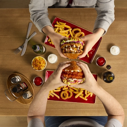 The hands of two people seated at a wooden table each holding a burger.  On teh table in front of each person are plates of fries and onion rings.