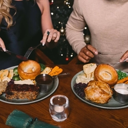 A close up view of two plates of Christmas dinner including Yorkshire pudding, roast potatoes, vegetables, stuffing, and pots of gravy and sauce. The plates sit on a wooden restaurant table along with drinks, a tealight, and a Christmas cracker; and two people sitting at the table are holding their knives and forks above the plates.