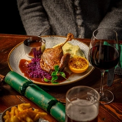 A plate of roasted duck served with mashed potatoes, red cabbage, and a slice of orange. The dish is garnished with green herbs and accompanied by a small metal cup of sauce. The plate is set on a wooden table with a glass of red wine, green napkin, fork, knife, and a Christmas cracker nearby. A person wearing a gray sweater is partially visible in the background.