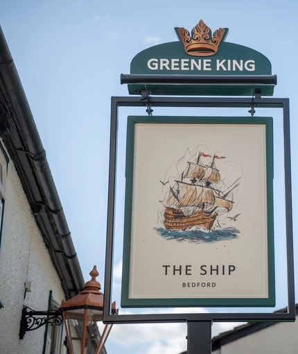 A close up view of the exterior pub sign of The Ship. The sign features an illustration of a large ship sailing on the sea and surrounded by seagulls, above the words "The Ship, Bedford", on a white background.