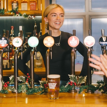 An image of a pint of Hazy Day sat on an interior bar with a person stood behind the bar with 2 customers stood in front.