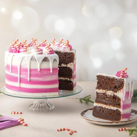 The Candy Cane Lane cake served on a glass cake stand which sits on a table scattered with pine tree sprigs and mini candy cane decorations. A slice has been cut out of the cake and served on a plate sitting on the table next to the cake stand.