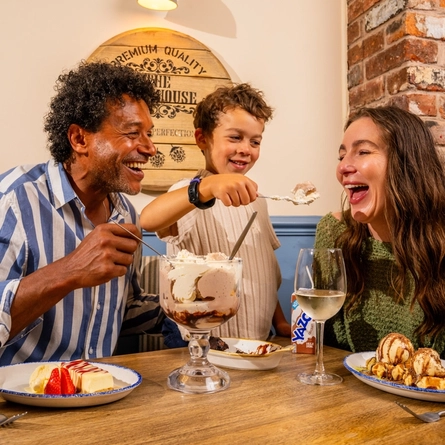 Two adults and a child are seated around a table in a pub, sharing a selection of desserts. The table features a large ice cream sundae, waffles topped with ice cream and sauce, cheesecake with strawberries, a chocolate dessert, a glass of white wine, a blue mug of coffee and a children's juice carton. Exposed brickwork, blue panelled walls and a wooden barrel-style wall sign create a warm and inviting pub setting.