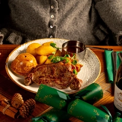 A person is seated at a wooden table. The table is decorated with festive decoration and is  holding a plated duo of beef dish.  Only the torso and arm is visible in the image. The person is holding a pint of Level Head.