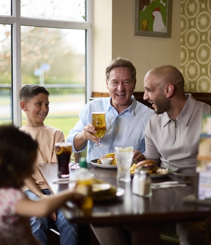 Two adults and two children sitting at a restaurant table in a Farmhouse Inns venue, laughing together. One of the adults is holding a glass of beer, and drinks and plates of food sit on the table in front of them.