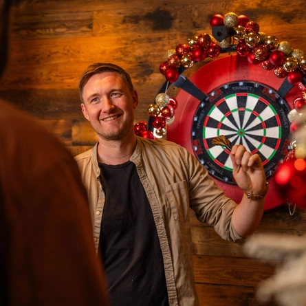 A person stands beside a electronic dartboard decorated with red, gold and silver Christmas baubles in a pub. Another person is visible in the foreground watching the game, while a snow-covered Christmas tree with red baubles frames the scene. Warm wooden panelling and festive decorations create a seasonal pub atmosphere.