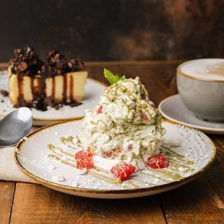 A close up view of a plate of Eton Mess and a plate of cheesecake. The plates sit on a cloth napkin on a wooden restaurant table, along with a spoon and a cup of coffee on a saucer.