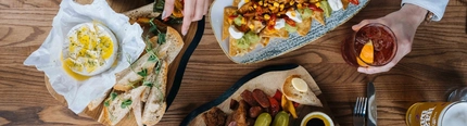 Overhead view of a wooden table with multiple sharing dishes and drinks. Two hands reach toward wooden boards topped with halved Scotch eggs with soft centres, gherkins, sliced sausage, fried chicken bites, roasted peppers, and thick slices of bread, served with small bowls of oil and dipping sauces. Nearby are a plate of loaded nachos topped with corn, sliced red chillies, guacamole, and sour cream, and a baked cheese round drizzled with oil, surrounded by bread slices and pickled vegetables.