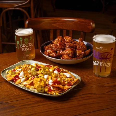 A Chicken Wing sharer served in a bowl, and a Nacho sharer served on a plate, sitting on a wooden restaurant table along with a glass of Level Head and a glass of Hazy Day.